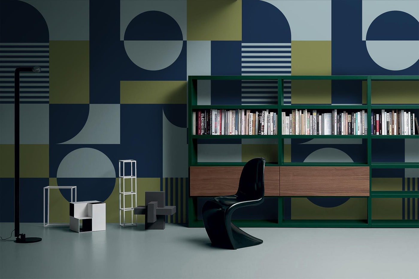 blue and green graphic wall mural on the wall of this living room. Shiny epoxy floor in grey. Green bookshelf with wooden doors. Black plastic chair