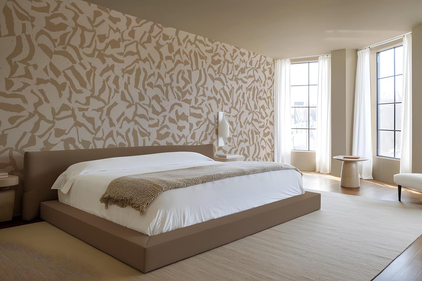 Graphic clay wall mural in sand on the wall of this neutral bedroom. Brown bed with white bedding and a beige throw. Light Beige rug on and light wooden floor