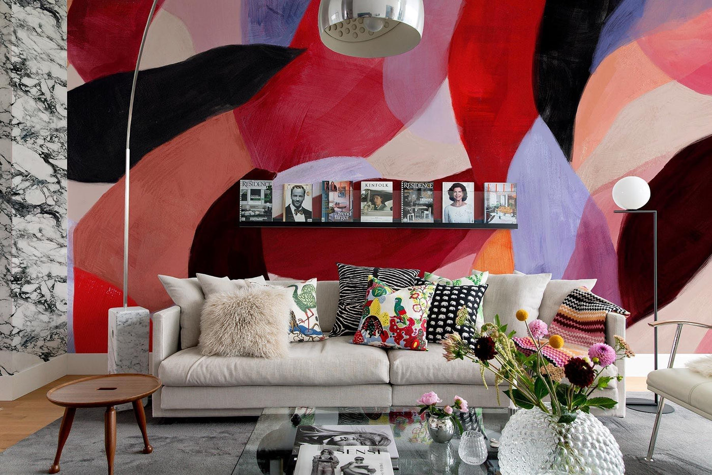 Mångfärgad konstnärlig väggvägg i röda toner i detta minimalistiska vardagsrum. Glastable. Arco lampa. träpall. Gray Rugg på trägolv. Grafisk kudde i soffan