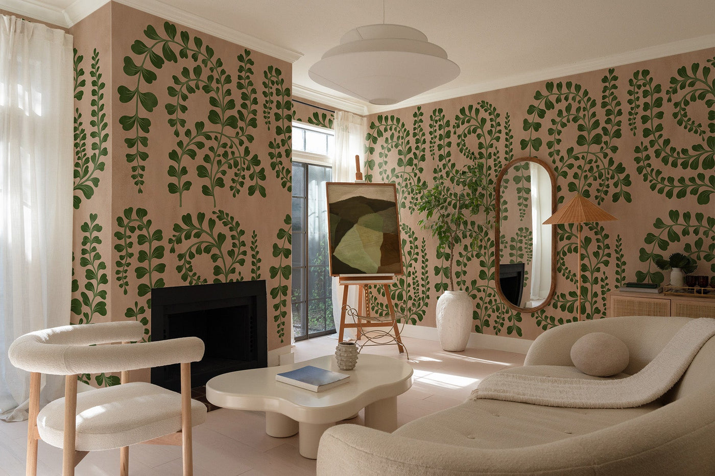 botanical wall mural in green and beige with leaves on the wall of this modern livingroom with white furniture. Curved white sofa. Curved white stool and a organic shaped coffe table. Oval mirror on the right wall with a rattan lamp