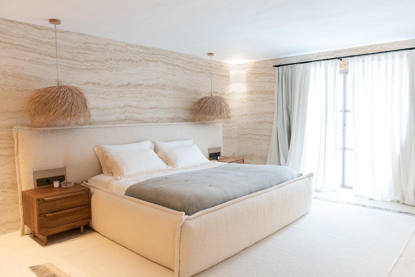Sandfärgad travertinvägg väggmålning på väggarna i detta moderna sovrum. Vitt golv. Lätt beige säng med vita sängkläder och ljusgrå filt. 