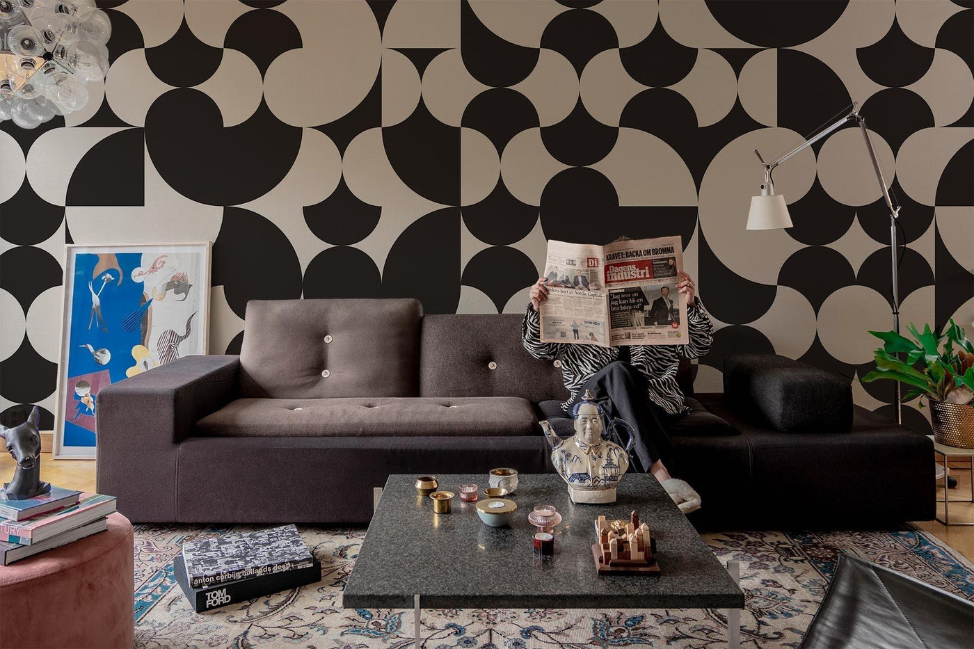 black and white geometric wall mural on the wall of this midcentury inspired living room. Black sofa. Vintage retro rug on the wooden floor. Black marble squared table and a vintage lamp