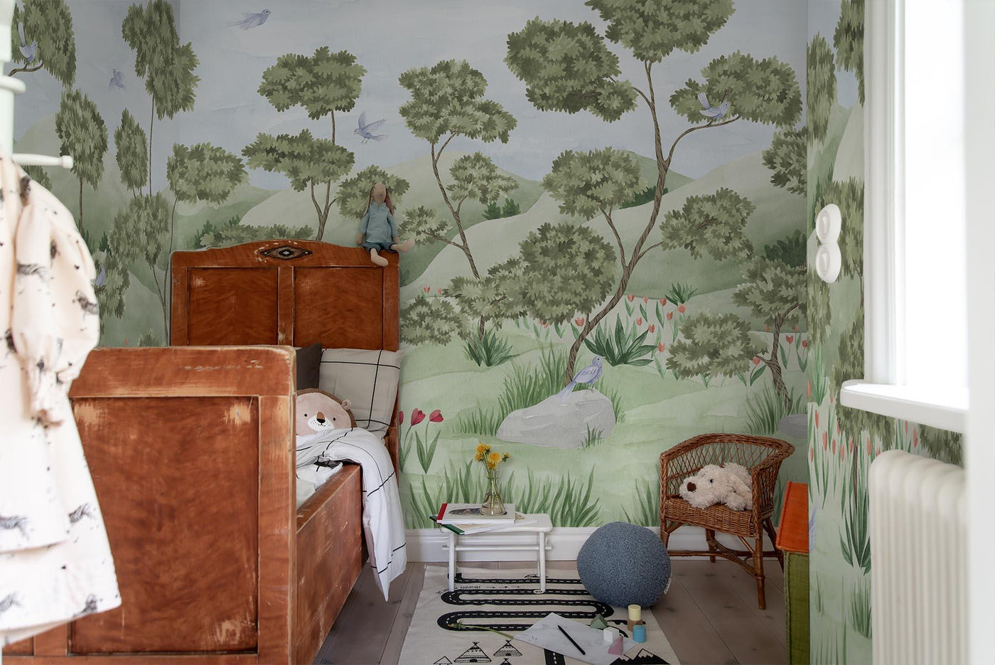 whimsical green wall mural of trees and birds in landscape on the walls of this kids room. Brown vintage kids bed in wood. Grey wooden floor. Toys on the floor